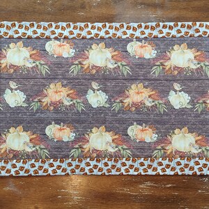 May include: A brown and white table runner with a fall theme. The runner features a repeating pattern of pumpkins, leaves, and birds on a woodgrain background. The runner has a brown and white border with a leaf pattern.