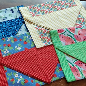May include: Four folded fabric placemats with different fruit designs. One placemat features blueberries, another strawberries, a third has watermelons, and the last one has a yellow and white checkered pattern.
