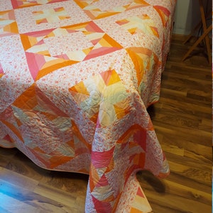 May include: A pink, orange, and white quilt with a floral pattern. The quilt is made up of squares and triangles, and it has a quilted design.