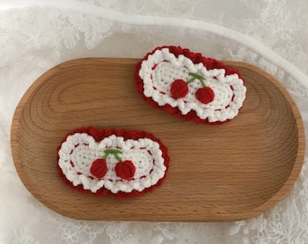 Puede incluir: Dos pinzas para el pelo de crochet rojo y blanco con un diseño de cereza. Las pinzas están sobre una superficie de madera.