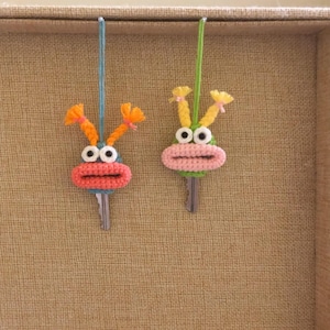 May include: Two crocheted keychains shaped like cartoon faces with big eyes, mouths, and antennas. The keychain on the left is pink, orange, and white. The keychain on the right is green, yellow, and white.