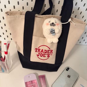 May include: A cream canvas tote bag with black handles and base, embroidered with the "Trader Joe's" logo. A plush seal keyring is attached. Other items include a pink hand sanitiser, a lip balm tube, a smartphone, and a small white box.