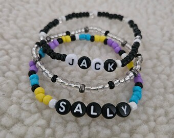 May include: Three beaded bracelets with the names "JACK" and "SALLY" spelled out in white letters on black beads. The bracelets are layered on top of each other, with the "JACK" bracelet on top and the "SALLY" bracelet on the bottom. The bracelets are made with black, white, yellow, blue, and silver beads.