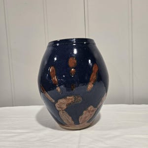 May include: A rounded ceramic vase with a glossy dark navy blue glaze. The surface features a prominent, textured handprint design in a light brown, earthy tone, revealing the raw clay texture where the glaze is absent on the palm and fingers.