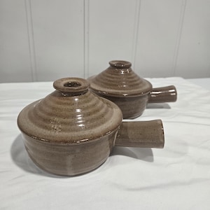 以下が含まれることがあります： 蓋とサイドハンドルが付いた、お揃いの茶色の陶器製スープボウル2個セット。各ボウルは、斑点のある釉薬仕上げ、蓋の上の円形のつまみ、丸みを帯びた本体が特徴です。ストーンウェア製の容器には、水平方向の畝のあるテクスチャーが施されています。