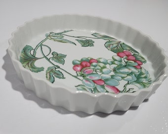 Könnte beinhalten: Weiße Keramik-Backform mit geriffeltem Rand. Die Form ist mit einem gemalten Design aus grünen Weinreben mit rosa und grünen Trauben verziert.