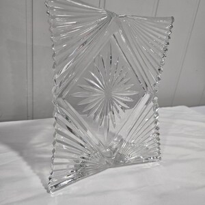 May include: A clear glass picture frame with a rectangular shape and a central diamond-shaped panel featuring a starburst pattern. The frame has radiating linear cuts extending to the edges and a decorative beaded border along the vertical sides.