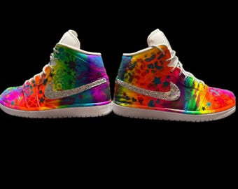 May include: Two high-top sneakers with a vibrant tie-dye design in shades of orange, yellow, green, blue, and purple. Each shoe features a silver glitter swoosh and white laces and soles. The shoes are decorated with star and floral patterns.