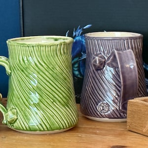 May include: Two ceramic mugs with textured, vertical lines. One is green, the other purple. Both have large handles and decorative elements. The mugs are on a wooden surface, with a notebook and a wooden box in the background.