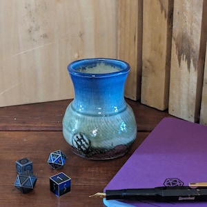 May include: A blue and green ceramic vase with a brown base, sitting on a wooden table with dice and a purple notebook with a pen.
