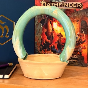 以下が含まれることがあります： 手作りのセラミックバスケット。ベースは淡いベージュで、取っ手はターコイズ色の曲線を描いています。木製の表面に置かれ、背景にはノートとペンが見えます。装飾用として使用される可能性が高いバスケットです。