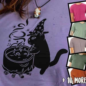 May include: A purple t-shirt featuring a black cat wearing a witch's hat and stirring a cauldron. The cat is surrounded by stars and steam. The text " + 10 MORE!" is in the bottom right corner of the image.
