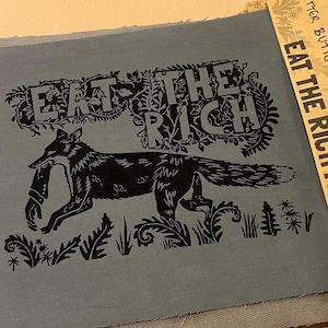 May include: A grey fabric patch featuring the text "EAT THE RICH" and a black fox illustration carrying a bundle. The patch is layered over denim fabric. A cardboard tag reads "CRITTER BUTTS EAT THE RICH PATCH".