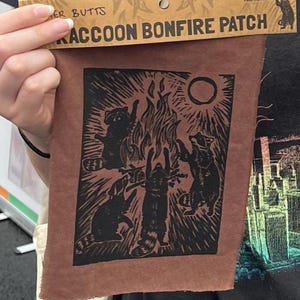 May include: A brown patch with a black illustration of raccoons around a bonfire, with the text "RACCOON BONFIRE PATCH". The patch is held up by a person. The patch has a rectangular shape.