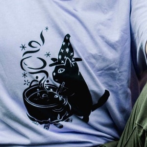 May include: A lavender t-shirt featuring a black graphic of a cat wearing a witch's hat and stirring a cauldron.