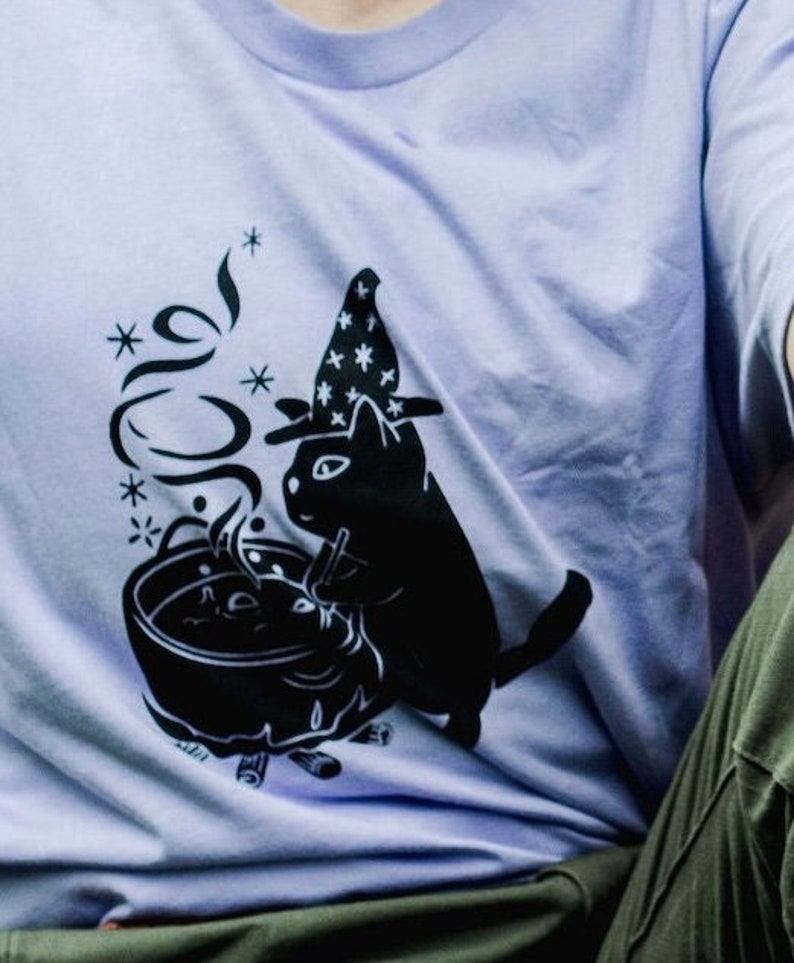 May include: A lavender t-shirt featuring a black graphic of a cat wearing a witch's hat and stirring a cauldron.