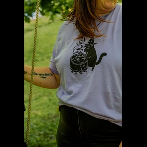 May include: A lavender t-shirt with a black graphic of a cat stirring a cauldron with a star on its head. The text on the shirt reads "There is only one thing for the dark."