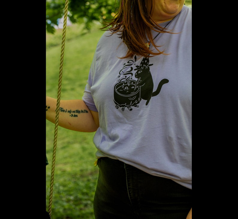 May include: A lavender t-shirt with a black graphic of a cat stirring a cauldron with a star on its head. The text on the shirt reads "There is only one thing for the dark."