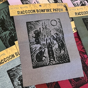 May include: A rectangular grey fabric patch featuring a black block print illustration of three raccoons standing around a campfire under a circular moon. The top header card is tan with the text CRITTER BUTTS and RACCOON BONFIRE PATCH.