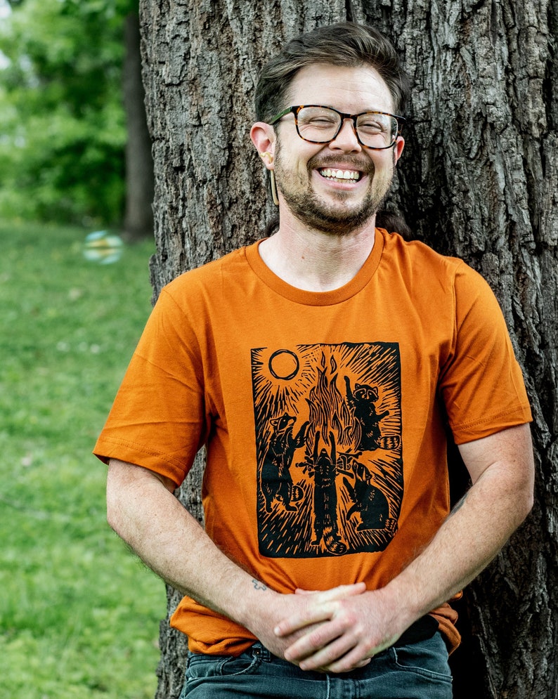 Pu&ograve; includere: Un uomo indossa una maglietta arancione con una stampa nera di procioni intorno a un fal&ograve;. La stampa include il testo "Campfire" e un sole sopra il fal&ograve;.