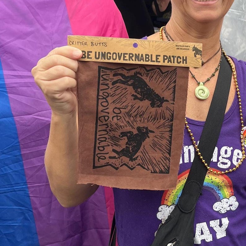 Może przedstawiać: Brązowa naszywka z napisem "BE UNGOVERNABLE PATCH" i "Critter Butts" wydrukowanym na g&oacute;rze. Naszywka zawiera dwie czarne ilustracje zwierząt i słowa "be ungovernable" w układzie pionowym.