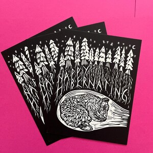 May include: Black and white illustration of a bear hibernating in a cave with trees and stars in the background. The text "Happy Hibernating" is written above the bear.