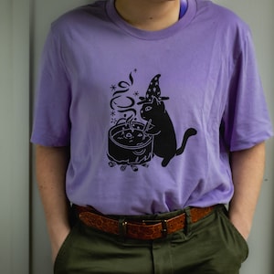 May include: A purple t-shirt featuring a black cat wearing a witch's hat and stirring a cauldron with a wand. The cat is standing next to a campfire with smoke rising from the cauldron.