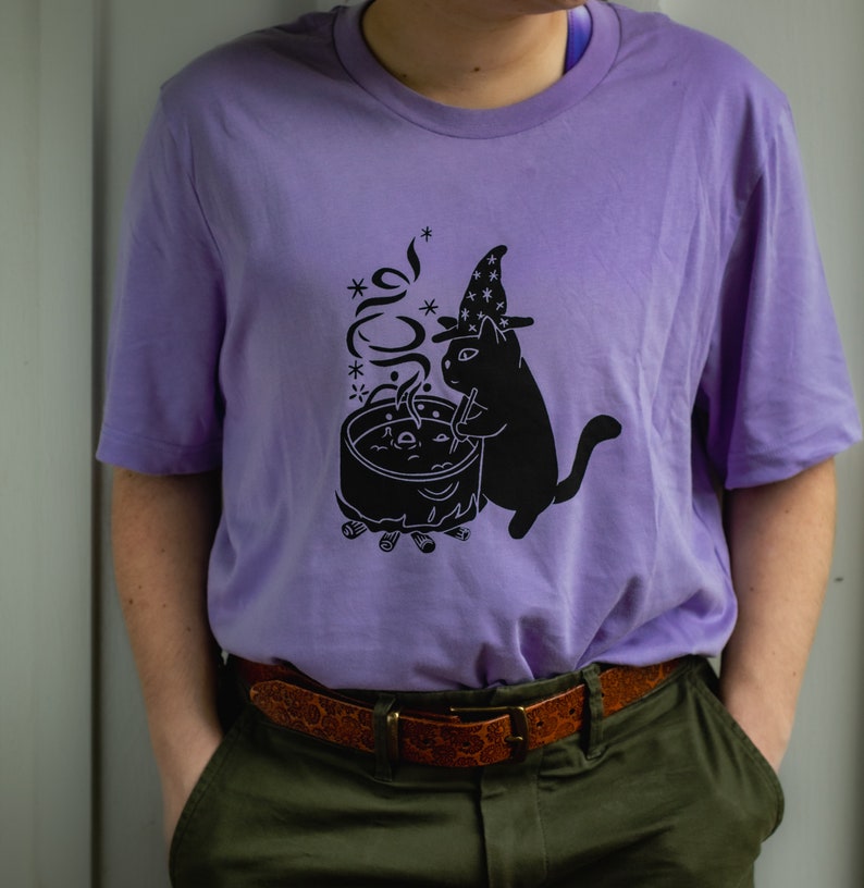 May include: A purple t-shirt featuring a black cat wearing a witch's hat and stirring a cauldron with a wand. The cat is standing next to a campfire with smoke rising from the cauldron.