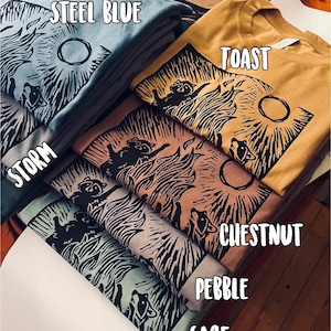 Pu&ograve; includere: Un mucchio di magliette con un design stampato di procione. Le magliette sono di vari colori, tra cui blu acciaio, tostato, tempesta, castano, ciottolo e salvia. Il testo "STEEL BLUE", "TOAST", "STORM", "CHESTNUT", "PEBBLE" e "SAGE" &egrave; visibile sulle magliette.