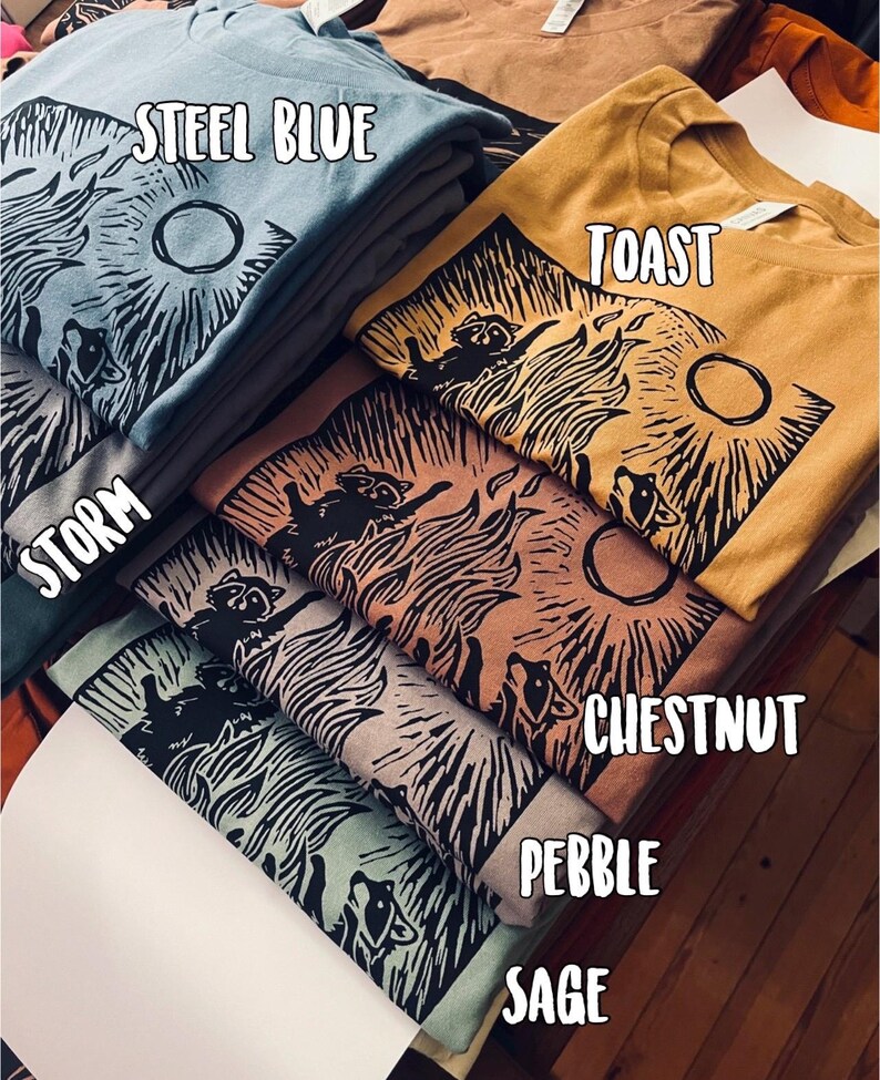 Pu&ograve; includere: Un mucchio di magliette con un design stampato di procione. Le magliette sono di vari colori, tra cui blu acciaio, tostato, tempesta, castano, ciottolo e salvia. Il testo "STEEL BLUE", "TOAST", "STORM", "CHESTNUT", "PEBBLE" e "SAGE" &egrave; visibile sulle magliette.