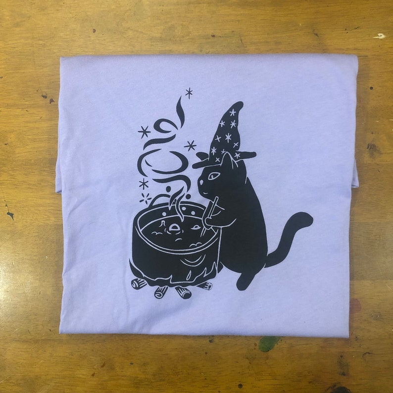 May include: A lavender t-shirt featuring a black illustration of a cat wearing a witch's hat and stirring a cauldron over a fire.
