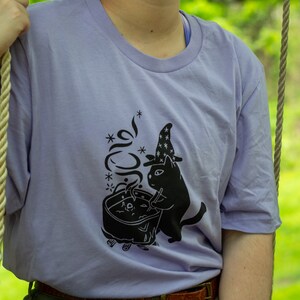 May include: A lavender t-shirt featuring a black graphic of a cat wearing a wizard hat and stirring a cauldron. The graphic includes the text "*meow*" with stars around it.