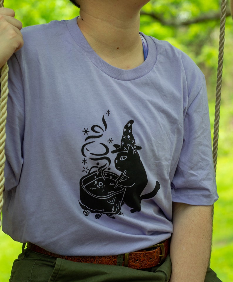 May include: A lavender t-shirt featuring a black graphic of a cat wearing a wizard hat and stirring a cauldron. The graphic includes the text "*meow*" with stars around it.
