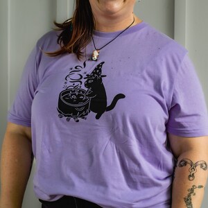 May include: A purple t-shirt featuring a black cat wearing a witch's hat and stirring a cauldron. The cat is surrounded by stars and smoke.