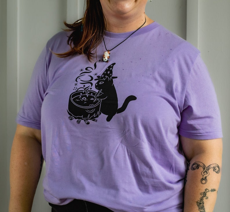 May include: A purple t-shirt featuring a black cat wearing a witch's hat and stirring a cauldron. The cat is surrounded by stars and smoke.