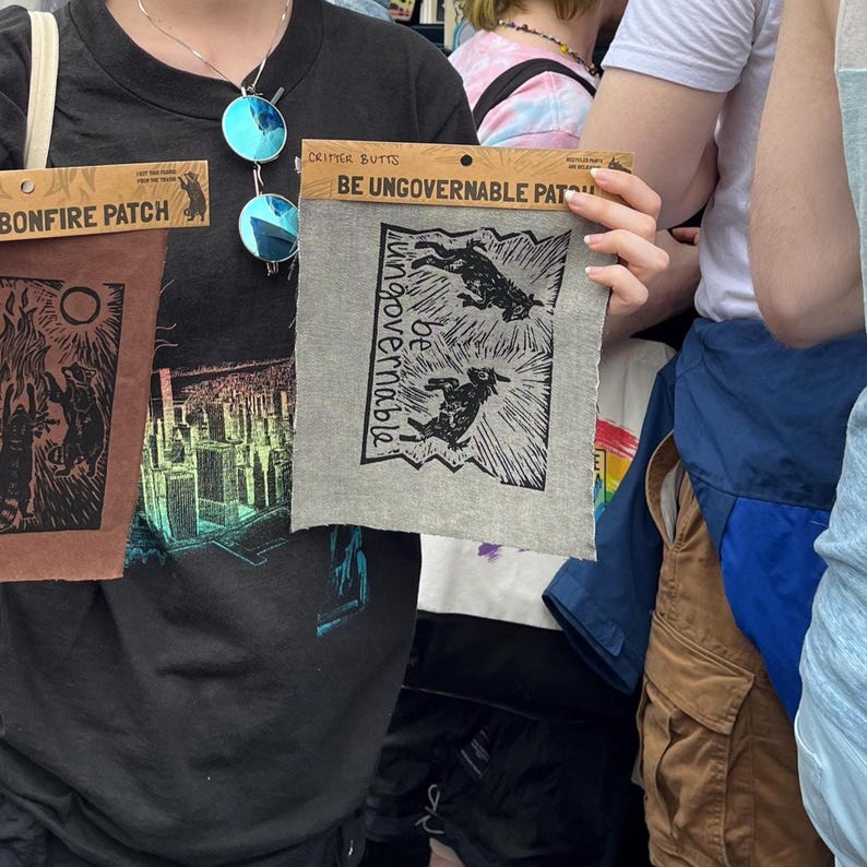 Może przedstawiać: Wyświetlane są trzy naszywki z tkaniny. Pierwsza naszywka jest brązowa z czarną ilustracją i napisem "BONFIRE PATCH". Druga naszywka jest szara z czarną ilustracją dw&oacute;ch zwierząt i napisem "BE UNGOVERNABLE PATCH". Trzecia naszywka jest szara z napisem "BE UNGOVERNABLE".