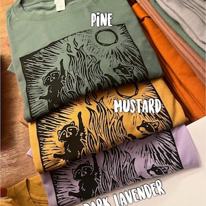 Pu&ograve; includere: Quattro t-shirt con un design grafico di procione in diversi colori: pino, senape, lavanda scuro e marrone chiaro. La grafica presenta procioni attorno a un fal&ograve; con la luna piena sullo sfondo.