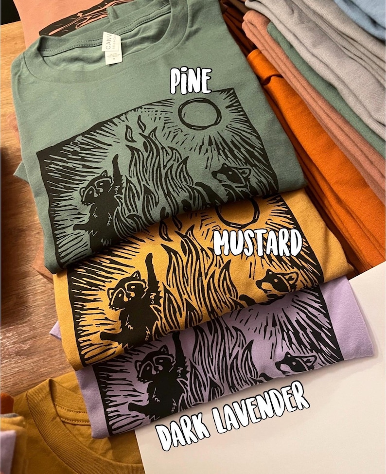 Pu&ograve; includere: Quattro t-shirt con un design grafico di procione in diversi colori: pino, senape, lavanda scuro e marrone chiaro. La grafica presenta procioni attorno a un fal&ograve; con la luna piena sullo sfondo.