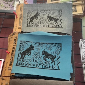 May include: A stack of square patches in shades of blue and gray, each featuring a black linocut print of two leaping goats and the words "be ungovernable." The patches are displayed on a wire rack with wooden tags that read "BE UNGOVERNABLE PATCH."