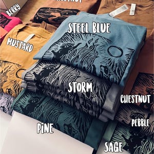 Pu&ograve; includere: Una pila di magliette di vari colori, tra cui salvia, ciottolo, castano, tempesta, pino, blu acciaio, senape e bacca. Ogni maglietta presenta una stampa grafica in bianco e nero di un procione.