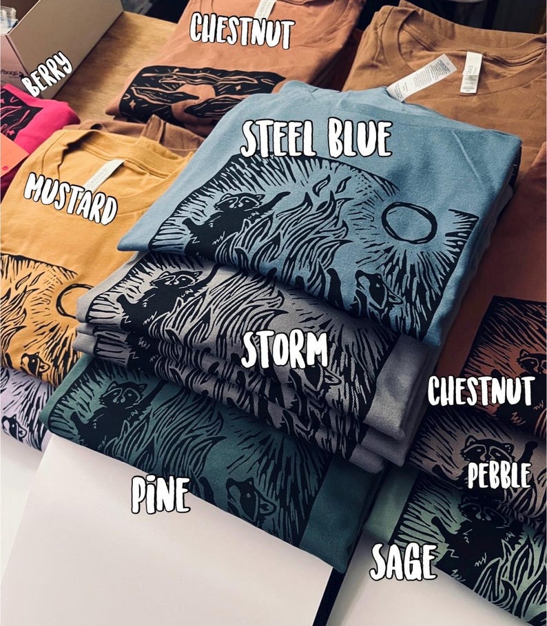 Pu&ograve; includere: Una pila di magliette di vari colori, tra cui salvia, ciottolo, castano, tempesta, pino, blu acciaio, senape e bacca. Ogni maglietta presenta una stampa grafica in bianco e nero di un procione.