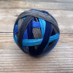 May include: A ball of yarn in shades of blue, gray, and black. The yarn is wound in a crisscross pattern, creating a textured appearance. The colors range from light blue to deep navy, with gray and black accents. The yarn is likely for knitting or crocheting.
