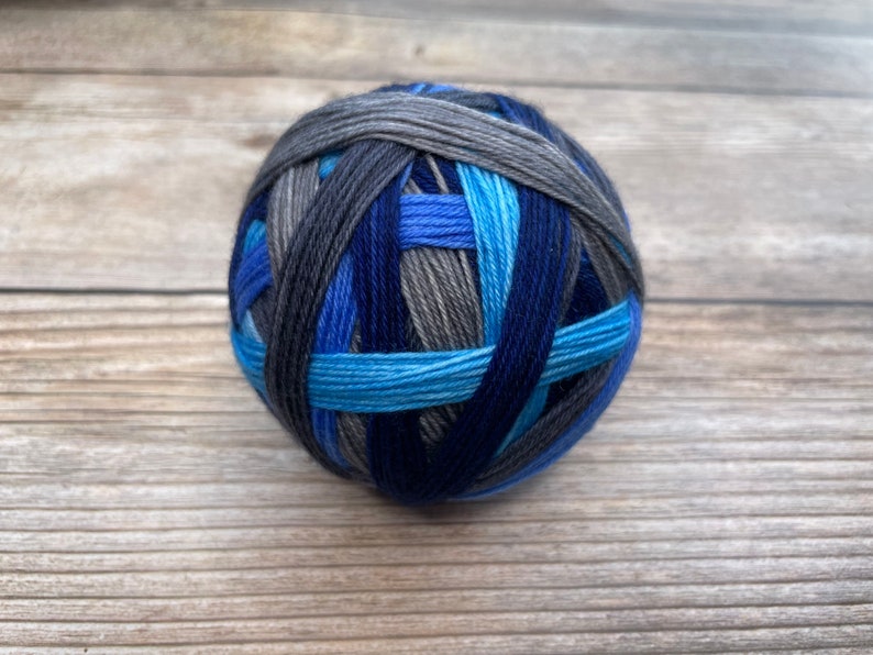 May include: A ball of yarn in shades of blue, gray, and black. The yarn is wound in a crisscross pattern, creating a textured appearance. The colors range from light blue to deep navy, with gray and black accents. The yarn is likely for knitting or crocheting.