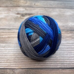 May include: A ball of yarn in shades of blue, gray, and black sits on a wooden surface. The yarn is tightly wound, with the colors blending together in a striped pattern. The yarn ball is a perfect sphere.