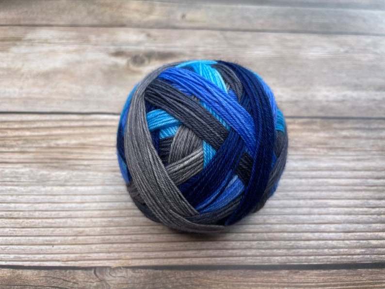 May include: A ball of yarn in shades of blue, gray, and black sits on a wooden surface. The yarn is tightly wound, with the colors blending together in a striped pattern. The yarn ball is a perfect sphere.