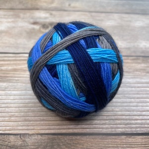 May include: A ball of yarn in shades of blue and gray, including navy, light blue, and charcoal. The yarn is tightly wound, showing the different colors woven together. Ideal for knitting or crocheting projects.