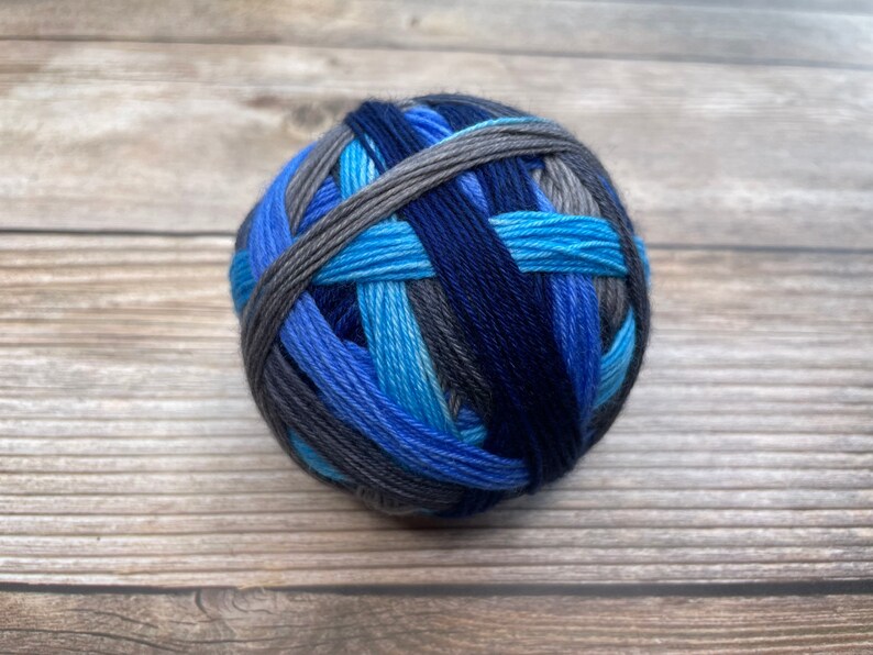 May include: A ball of yarn in shades of blue and gray, including navy, light blue, and charcoal. The yarn is tightly wound, showing the different colors woven together. Ideal for knitting or crocheting projects.