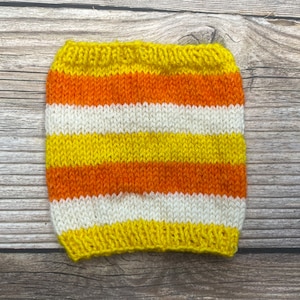 Puede incluir: Un accesorio tejido a mano al estilo de los caramelos de maíz. El accesorio presenta rayas horizontales amarillas, naranjas y blancas. El artículo está hecho de hilo y está diseñado para adaptarse a un recipiente para bebidas.