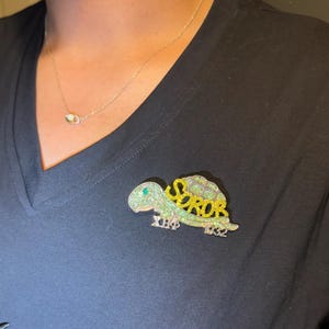 May include: A glittery turtle-shaped brooch with the word "SOROB" in yellow and green. The brooch also features the Greek letters "XΦ" and the number "1932". The brooch is pinned to a black v-neck shirt.