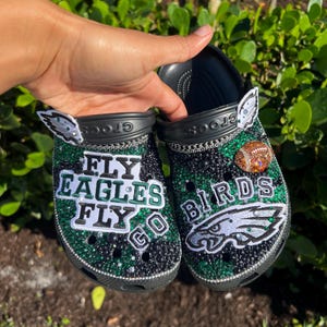 Può includere: Un paio di zoccoli neri decorati con strass verdi e neri, con toppe che riportano le scritte FLY EAGLES FLY GO e BIRDS, un logo a forma di aquila, un ciondolo a forma di pallone da football e catenelle argentate sui cinturini.